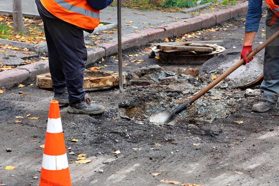 Two construction workers uses shovels to dig up a sewer pipe for sewer maintenance and stormwater repair.